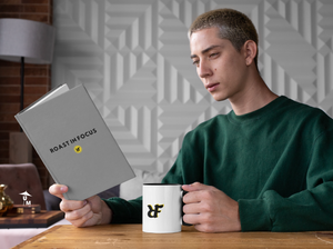 Man reading book at office desk with white ceramic "RIF" Roast In Focus Logo Coffee mug. Black & yellow design. 11oz cup with colored rim. Green sweatshirt cool guy.