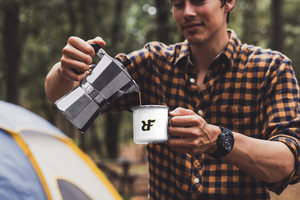 Man camping in the woods with Gshock watch pouring coffee in his white enamel "RIF" Roast In Focus Logo Coffee mug. Black & yellow design. 13oz cup, durable, lightweight bring into nature.